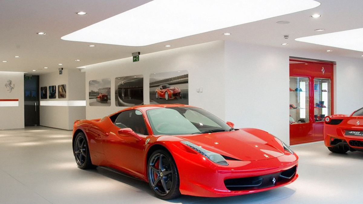 Red sports car inside a modern Ferrari showroom illuminated by large recessed white ceiling lightboxes.