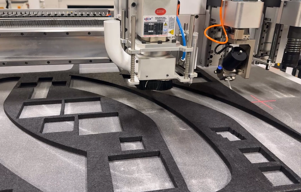 Automated CNC router head precision cutting geometric shapes into a dark acoustic panel on a flatbed table.