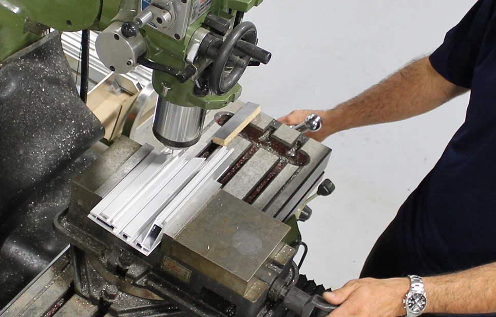 Machinist operating a milling machine to precision cut aluminum extrusions clamped in a heavy-duty vise.
